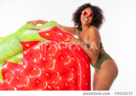 Pretty african female stands in swimsuit with rubber beach strawberry mattress and smiles isolated Pretty african female stands in swimsuit with rubber beach strawberry mattress and smiles isolated 59529237