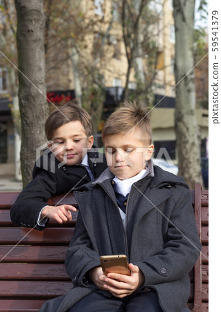 Two schoolchildren are watching an interesting video on a smartphone and smiling while sitting on a Two schoolchildren are watching an interesting video on a smartphone and smiling while sitting on a 59541379
