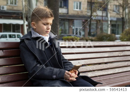 A school-aged boy sits on a bench in a park in a beautiful business coat and looks at something on 59541413