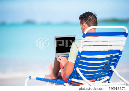 Young man with laptop on tropical caribbean beach. Man sitting on the sunbed with computer and 59548871