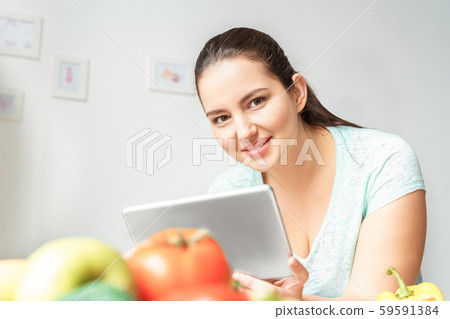 Body Care. Chubby girl leaning on table with vegetables in kitchen doing online shopping on digital Body Care. Chubby girl leaning on table with vegetables in kitchen doing online shopping on digital 59591384