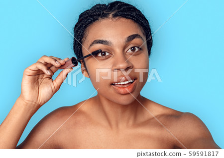 Beauty Concept. Young african woman isolated on blue applying mascara smiling concentrated close-up Beauty Concept. Young african woman isolated on blue applying mascara smiling concentrated close-up 59591817