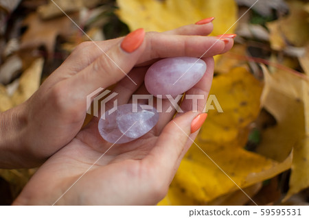 Female hands with orange manicure holding two yoni eggs made from pink quartz and transparent violet 59595531