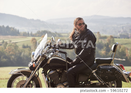 Bearded biker with long hair in black leather jacket and sunglasses sitting on modern motorcycle. 59610272