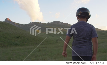 a boy in a motorcycle helmet walks on a hillside against the backdrop of mountains backlit by sunset a boy in a motorcycle helmet walks on a hillside against the backdrop of mountains backlit by sunset 59617731