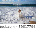 A young man in the water on a sandy beach walking into the foamy wave 59617794