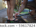 Father is washing child's feet with a water from a garden hose 59617824