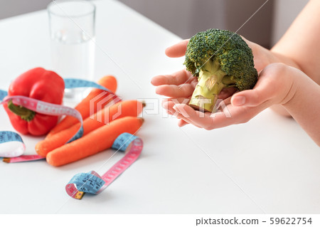 Body Care. Chubby girl sitting at kitchen table with vegetables and tape measure holding broccoli Body Care. Chubby girl sitting at kitchen table with vegetables and tape measure holding broccoli 59622754