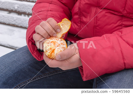 A man peels a tangerine while sitting on a bench in a winter park 59645560