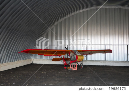 Very small single-place ultralight glider stands in the corner of a metal hangar 59686173
