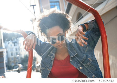 Outdoors Leisure. Young stylish african girl in denim jacket and sunglasses standing leaning on bar 59706858