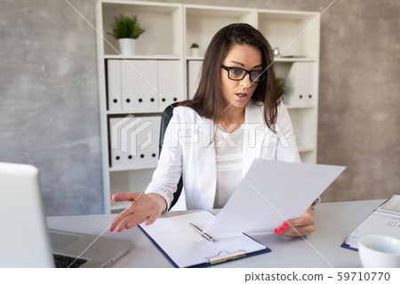 The dark-haired businesswoman looks in surprise at what is written on a piece of paper in her hands The dark-haired businesswoman looks in surprise at what is written on a piece of paper in her hands 59710770