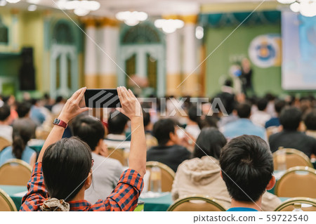 Closeup woman audience hand holding smart mobile phone for taking photo or doing Live streaming to Asian Speaker with casual suit on the stage present the screen in conference hall or seminar meeting 59722540