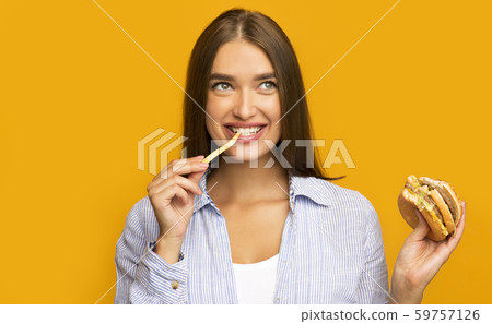 Happy Girl Eating Burger And French Fries Standing, Yellow Background Happy Girl Eating Burger And French Fries Standing, Yellow Background 59757126