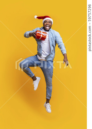 Black guy holding christmas gift and jumping over yellow background Black guy holding christmas gift and jumping over yellow background 59757296