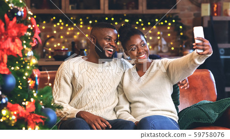 Cheerful afro couple taking selfie near Christmas tree at home 59758076