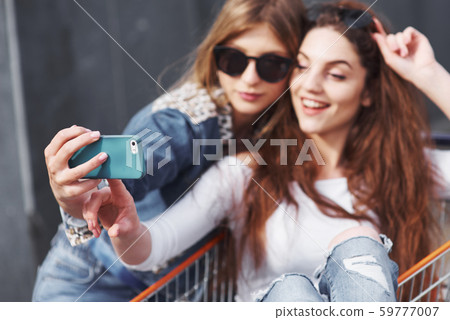 Two young happy women in sunglasses having fun shopping trolley race outdoors. Two young happy women in sunglasses having fun shopping trolley race outdoors. 59777007
