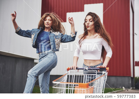 Two young happy women having fun shopping trolley race outdoors. 59777383