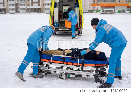 Two paramedics in blue workwear fixing unconscious man with belts on stretcher 59810011