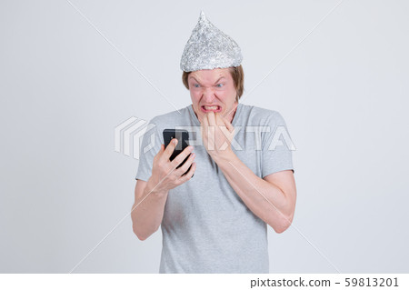 Stressed young man with tin foil hat using phone and looking scared 59813201