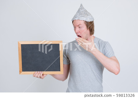Young man with tin foil hat holding blackboard and looking shocked Young man with tin foil hat holding blackboard and looking shocked 59813326