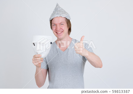 Happy young man with tinfoil hat holding white mask and giving thumbs up Happy young man with tinfoil hat holding white mask and giving thumbs up 59813387