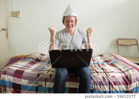 Happy young man with tin foil hat using laptop and getting good news in the bedroom 59813577