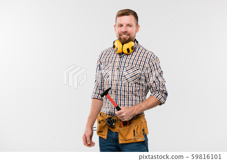 Bearded smiling technician with protective earphones, hammer and tool belt 59816101