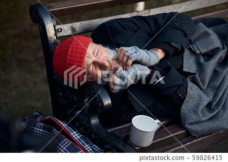 View of frozen old man lying on bench in city park in cold weather View of frozen old man lying on bench in city park in cold weather 59826415