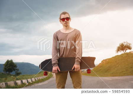 Stylish young man standing along a winding mountain road with a skate or longboard on his shoulder Stylish young man standing along a winding mountain road with a skate or longboard on his shoulder 59854593