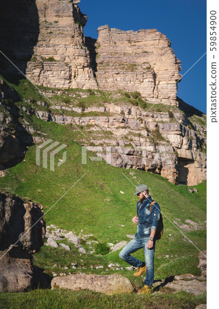 Stylish young man with a long beard in a jeans suit, sunglasses and a cap with a backpack stands on Stylish young man with a long beard in a jeans suit, sunglasses and a cap with a backpack stands on 59854900