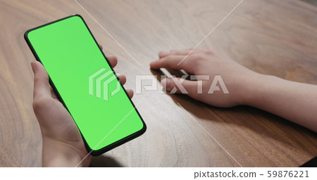 Young woman sitting at a table and using a smartphone with vertical green screen 59876221