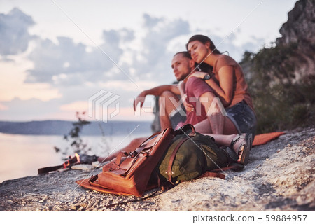 Hugging couple with backpack sitting near the fire on top of mountain enjoying view coast a river or 59884957