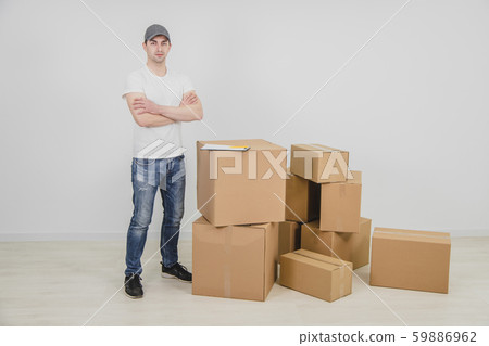 Strong young delivery man standing with his hands folded, near a stack of cardboard packages. Strong young delivery man standing with his hands folded, near a stack of cardboard packages. 59886962