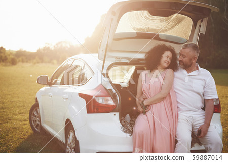 Grandpa and grandmother sit together on the nature near the car. They make a selfie photo and their 59887014