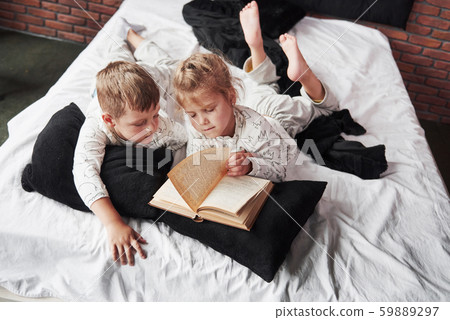 Two children lie on a large bed and read an interesting book. They are dressed in the same pajamas Two children lie on a large bed and read an interesting book. They are dressed in the same pajamas 59889297