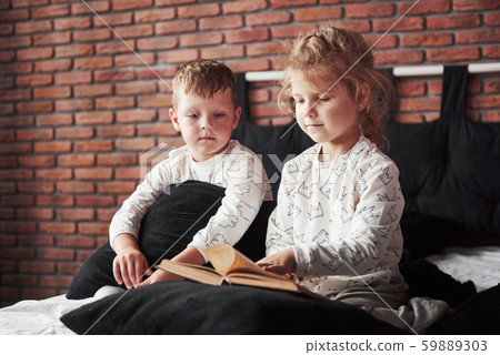 Two children lie on a large bed and read an interesting book. They are dressed in the same pajamas 59889303