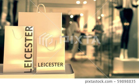 Paper shopping bags with Leicester caption against blurred store entrance. Retail in the United 59898835