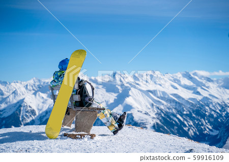 Photo from back of athlete man sitting on bench next to snowboard in ski resort. 59916109