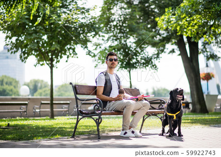 Young blind man with white cane and guide dog sitting in park in city. 59922743