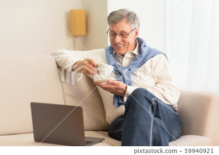 Elderly Man Using Laptop Having Coffee Sitting On Couch Indoor Elderly Man Using Laptop Having Coffee Sitting On Couch Indoor 59940921