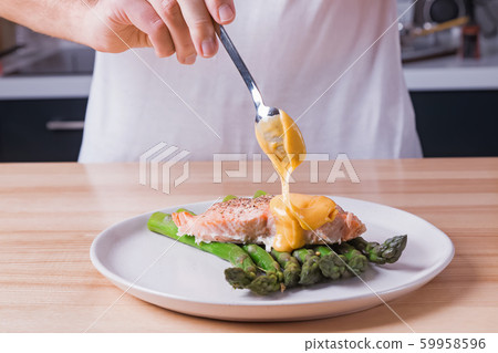 Man's hands pouring hollandaise sauce on top of delicious baked salmon with steamed green asparagus Man's hands pouring hollandaise sauce on top of delicious baked salmon with steamed green asparagus 59958596