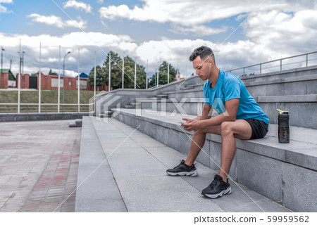 male athlete sits, holds mobile phone hands, listens music headphones, rest after summer workout in city, background cloud step. Active youth lifestyle, fitness nature. Free space motivation text. male athlete sits, holds mobile phone hands, listens music headphones, rest after summer workout in city, background cloud step. Active youth lifestyle, fitness nature. Free space motivation text. 59959562