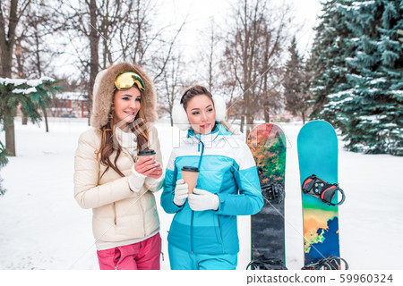 Two girls girlfriends winter park resort. Relaxing, talking warming up hot drinks, cup coffee tea. Emotions pleasure comfort. Sports overalls, background snow trees snowboards. Two girls girlfriends winter park resort. Relaxing, talking warming up hot drinks, cup coffee tea. Emotions pleasure comfort. Sports overalls, background snow trees snowboards. 59960324