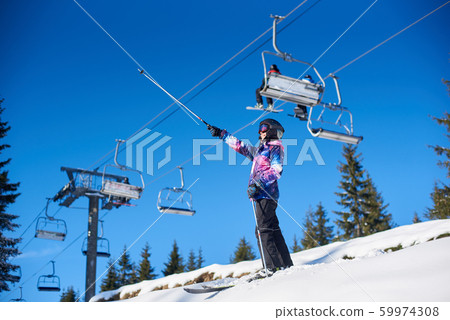 Happy woman skier standing near ski lift on snow-covered mountain slope. Sunny day during winter vacation. General view. Happy woman skier standing near ski lift on snow-covered mountain slope. Sunny day during winter vacation. General view. 59974308