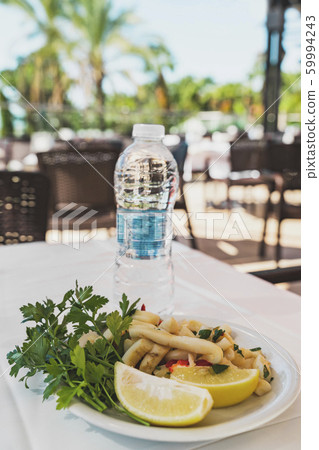 Squid in a white plate served with lemon and parsley, nearby is bottle with mineral water. The dish is on the table with a white tablecloth in a summer cafe outdoors. Healthy lifestyle and natural foo 59994243