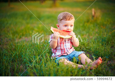 Happy child sitting on green grass and eating watermelon outdoors in spring park against natural background 59994672