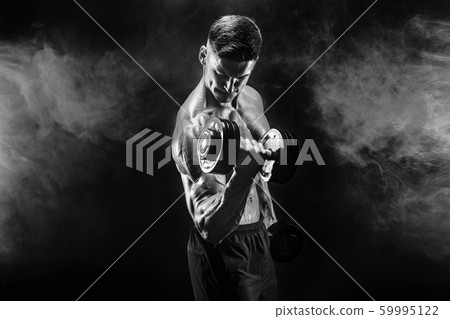 Handsome bodybuilder doing exercise with dumbbell. Studio shot. Black and white photo. Smoke Handsome bodybuilder doing exercise with dumbbell. Studio shot. Black and white photo. Smoke 59995122