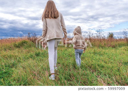 Family mom woman with a little boy 4-5 years old on weekends walk in the park field, background green grass. Warm casual wear, hooded sweater. Caring support love and parenting hold hands. Back view. 59997009