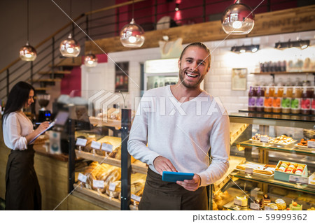 Bearded businessman holding tablet smiling while owning bakery with wife Bearded businessman holding tablet smiling while owning bakery with wife 59999362